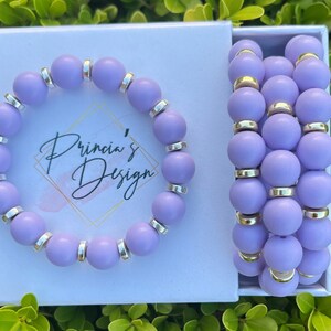 May include: A set of three lavender-colored beaded bracelets with silver accents. The bracelets are packaged in a white box with the text "Princia's Design" printed on the front.