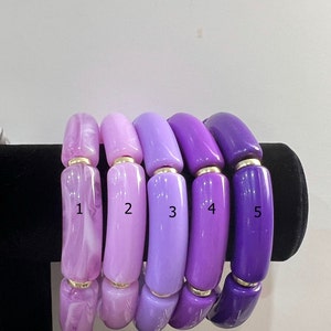 May include: Five purple and lavender bangle bracelets with gold accents. The bracelets are arranged in a gradient from light to dark purple.