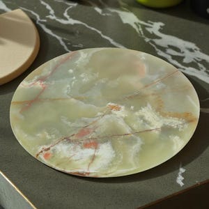 May include: A round, light green onyx coaster with red and white veining sits on a dark green marble-patterned surface. A beige half-moon coaster is to the left. Other items include a green water bottle and a white container.