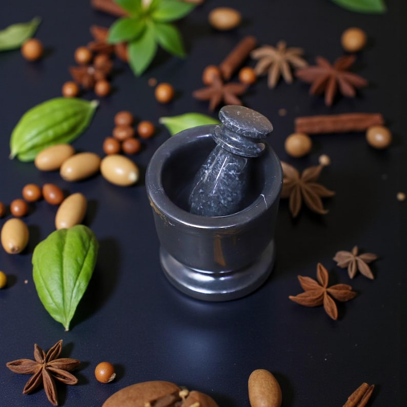 A black mortar and pestle set is surrounded by various spices and herbs on a black surface. Star anise, cinnamon sticks, and green leaves are scattered around the mortar, creating a natural, earthy aesthetic.