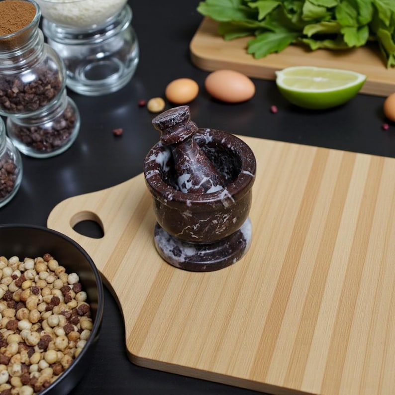 A red marble mortar and pestle set with white veining sits on a bamboo cutting board. The mortar is round with a flared rim, and the pestle is cylindrical. Ingredients and other kitchen items are in the background.
