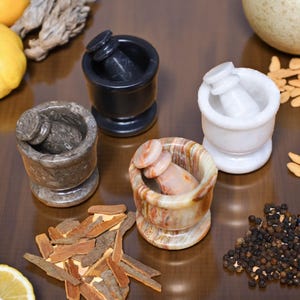 Four stone mortar and pestle sets in black, white, gray, and multicolored patterns. The mortars are cylindrical with a rounded base, and the pestles have a rounded handle. Spices and ingredients are scattered around the mortars.