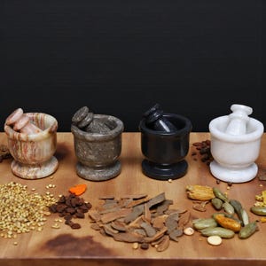 Four stone mortar and pestle sets in various colors: green , gray, black, and white. Spices and herbs are arranged on a wooden surface in front of the mortars. The background is black.