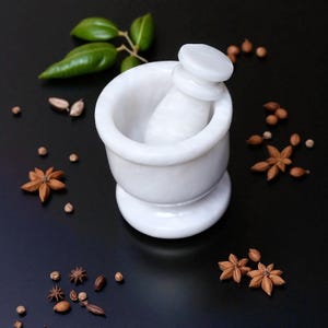 A white marble mortar and pestle set. The mortar has a wide bowl and a rounded base. The pestle is cylindrical with a flat top. The set is surrounded by star anise, peppercorns, and green leaves on a black surface.