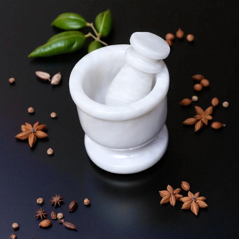 A white marble mortar and pestle set. The mortar has a wide bowl and a rounded base. The pestle is cylindrical with a flat top. The set is surrounded by star anise, peppercorns, and green leaves on a black surface.