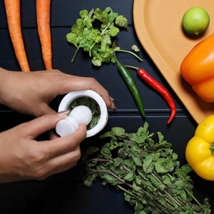 A white mortar and pestle is used to grind herbs, surrounded by fresh ingredients. The image includes carrots, peppers, a lime, and chili peppers on a dark surface, suggesting a culinary theme.