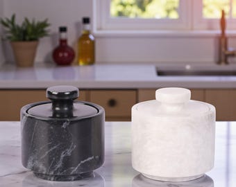 May include: Two marble canisters with lids. One is black with white veining, the other is white. Both have a cylindrical shape with a small, round knob on top of the lid. They sit on a marble countertop in a kitchen setting.