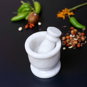 A white marble mortar and pestle set. The mortar has a rounded bowl and a pedestal base. The pestle is cylindrical with a rounded top. Green peppers, spices, and a coconut are in the background.