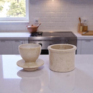 Op de afbeelding: Twee beige stenen keukenaccessoires staan op een wit aanrechtblad. Eén is een kleine, ronde container met een brede opening. De andere is een kopvormig object op een ronde basis. Op de achtergrond een fornuis, raam en witte tegels.