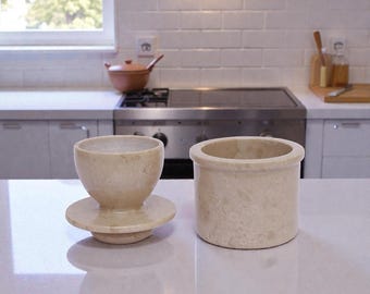 Könnte beinhalten: Zwei Küchenaccessoires aus beigem Stein stehen auf einer weißen Arbeitsplatte. Eines ist ein kleiner, runder Behälter mit einer weiten Öffnung. Das andere ist ein becherförmiges Objekt auf einem runden Sockel. Im Hintergrund befinden sich ein Herd, ein Fenster und weiße Fliesen.