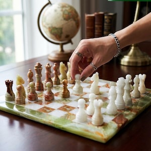 May include: A chess set with a green and white marble board. The chess pieces are in shades of brown, beige, and white. A hand is moving a white pawn on the board. A globe and books are in the background.