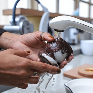 Puede incluir: Un tap&oacute;n de botella de piedra marr&oacute;n y blanca se sostiene bajo agua corriente. El tap&oacute;n tiene forma redondeada con una base plana y circular. El agua fluye de un grifo blanco.