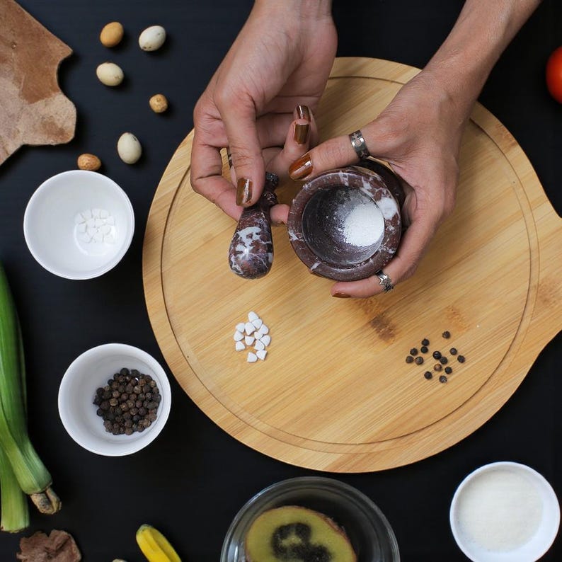A dark red and white speckled mortar and pestle set with white salt inside the mortar. The set is on a wooden cutting board with various ingredients, including peppercorns, white bowls, and a kiwi slice. The background is black.