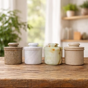 Op de afbeelding: Vier stenen potten met deksels, elk in een andere kleur en textuur, staan op een houten oppervlak. De potten variëren van lichtbeige tot gemarmerd wit en groen-gele onyx. De deksels zijn rond en passen goed op de potten.