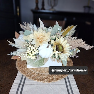 May include: A white box filled with a fall floral arrangement featuring white pumpkins, sunflowers, and dried grasses. The arrangement is sitting on a woven jute round placemat on a white and gray striped table runner. The text "@juniper_farmhouse" is visible in the bottom right corner of the image.