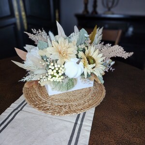 May include: A white ceramic planter filled with a floral arrangement of white and cream flowers, dried grasses, and white berries. The planter is sitting on a woven jute placemat on a brown table with a white and black striped runner.