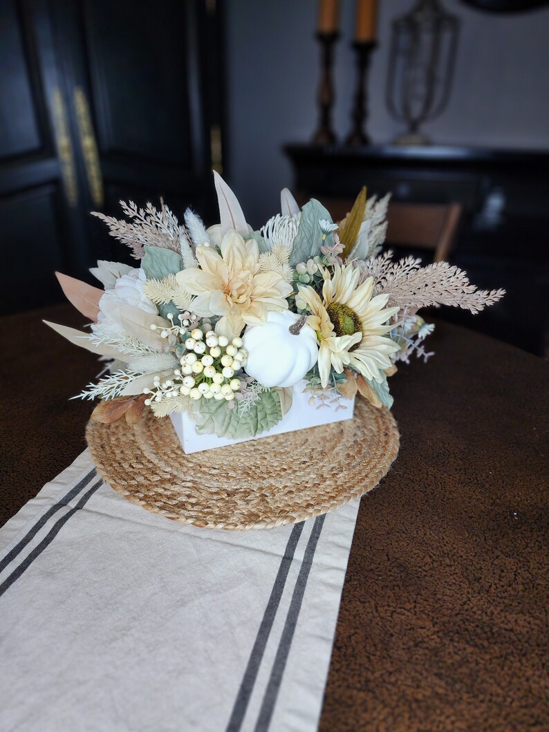 May include: A white ceramic planter filled with a floral arrangement of white and cream flowers, dried grasses, and white berries. The planter is sitting on a woven jute placemat on a brown table with a white and black striped runner.