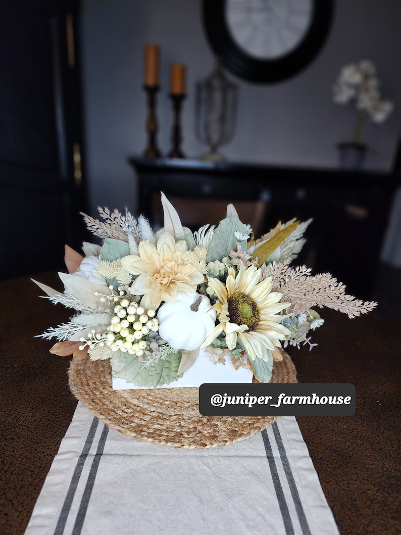 May include: A white box filled with a fall floral arrangement featuring white pumpkins, sunflowers, and dried grasses. The arrangement is sitting on a woven jute round placemat on a white and gray striped table runner. The text "@juniper_farmhouse" is visible in the bottom right corner of the image.