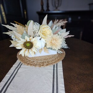 May include: A white ceramic planter with a faux floral arrangement featuring sunflowers, dahlias, and dried grasses. The planter is sitting on a jute placemat and a white table runner with black stripes.