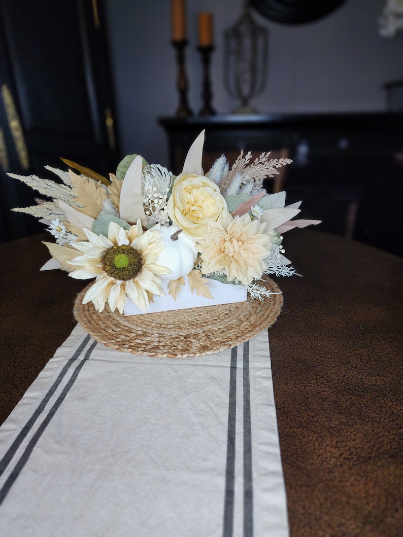 May include: A white ceramic planter with a faux floral arrangement featuring sunflowers, dahlias, and dried grasses. The planter is sitting on a jute placemat and a white table runner with black stripes.