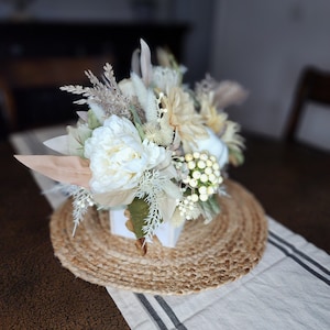 May include: A white and cream floral arrangement in a white square vase, sitting on a round woven jute placemat. The arrangement includes white peonies, dried grasses, and white berries.