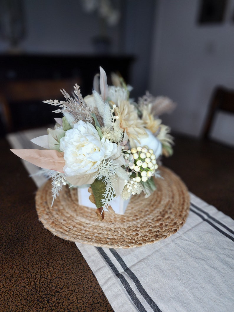 May include: A white and cream floral arrangement in a white square vase, sitting on a round woven jute placemat. The arrangement includes white peonies, dried grasses, and white berries.