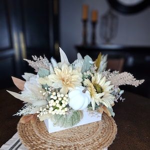 May include: A white ceramic pumpkin sits in a white box surrounded by a variety of dried and faux flowers in shades of cream, beige, and brown. The arrangement is on a woven jute placemat.