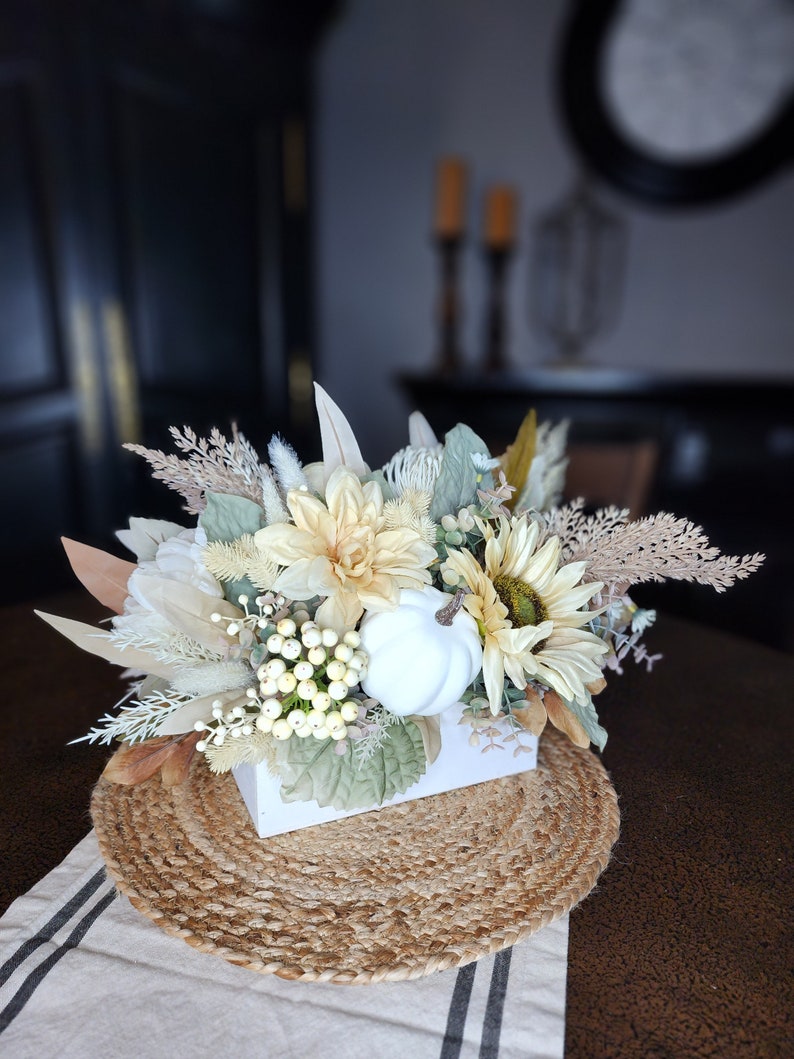 May include: A white ceramic pumpkin sits in a white box surrounded by a variety of dried and faux flowers in shades of cream, beige, and brown. The arrangement is on a woven jute placemat.