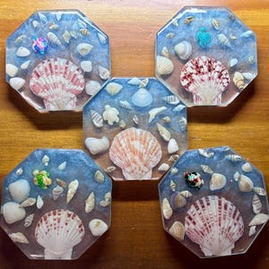 May include: Five octagonal resin coasters with a beach theme. Each coaster features a large seashell, smaller shells, and a miniature turtle. The resin is a translucent blue, and the seashells are white and pink.