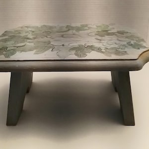 May include: A small, wooden stool with a light blue paint finish. The top of the stool is decorated with a leafy design in shades of green and white.