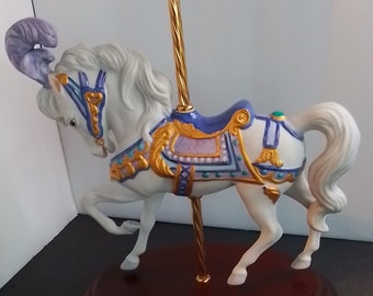 May include: A white porcelain carousel horse with a purple and blue saddle and gold accents. The horse is standing on a wooden base with a gold pole.