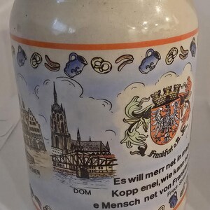 Puede incluir: Una jarra de cerveza de cerámica beige con una ilustración detallada de una escena de la ciudad, un escudo de armas y texto. La jarra presenta un borde decorativo con pretzels y jarras azules.