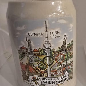 May include: Cream-colored ceramic beer stein with a colorful illustration of the Munich Olympic Park, including the Olympic Tower (290m) and the Olympic rings. The stein has a rounded shape and a handle.