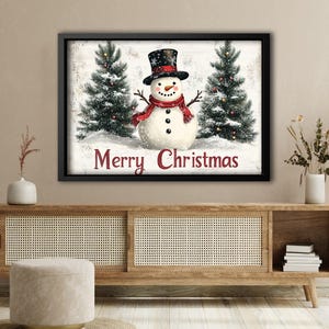 K&ouml;nnte beinhalten: Ein gerahmter Weihnachtsdruck mit einem Schneemann mit rotem Schal und schwarzem Zylinder, der zwischen zwei schneebedeckten Kiefern steht. Der Text "Merry Christmas" ist in Rot unter dem Schneemann geschrieben.