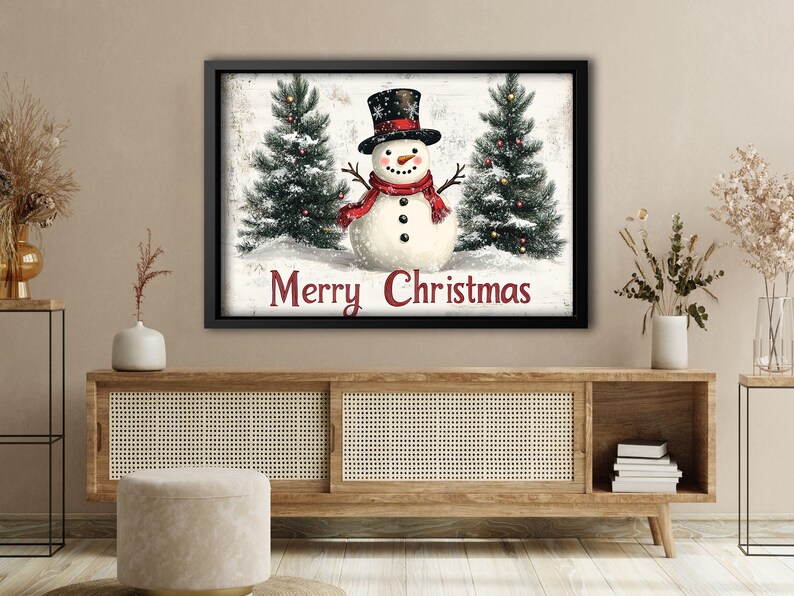 K&ouml;nnte beinhalten: Ein gerahmter Weihnachtsdruck mit einem Schneemann mit rotem Schal und schwarzem Zylinder, der zwischen zwei schneebedeckten Kiefern steht. Der Text "Merry Christmas" ist in Rot unter dem Schneemann geschrieben.