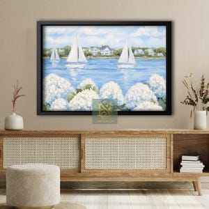 May include: A framed painting depicts sailboats on a blue sea, with white flowers in the foreground and houses in the background. The artwork is encased in a black frame, creating a coastal theme.