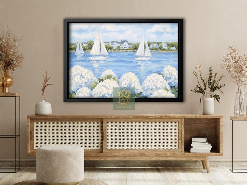 May include: A framed painting depicts sailboats on a blue sea, with white flowers in the foreground and houses in the background. The artwork is encased in a black frame, creating a coastal theme.