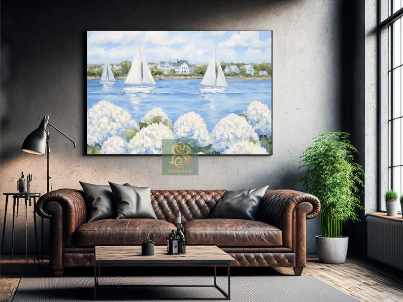 May include: A framed landscape painting depicts sailboats on a blue sea, with white hydrangeas in the foreground. The sky is blue with clouds, and houses are visible in the distance. The painting is displayed above a brown leather sofa.
