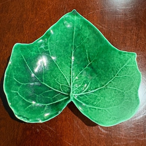 May include: A glossy, vibrant green ceramic dish shaped like a large leaf. The surface features a detailed, raised vein pattern that radiates from the center stem, creating a realistic botanical texture with a smooth, reflective finish.