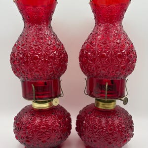 May include: A pair of matching red glass oil lamps featuring a textured starburst and hobnail pattern. Each lamp consists of a rounded base and a bulbous chimney with a ruffled top edge, connected by a brass-colored metal burner mechanism.