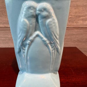 May include: A light blue ceramic vase featuring a raised relief design of two parakeets perched together on a branch. The vase has a flared top opening and a rectangular base, with a smooth, glossy finish covering the entire exterior surface.