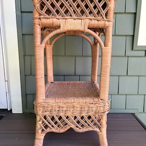May include: A tall, light brown wicker plant stand featuring a square top surface, a lower shelf, and four vertical legs. The structure is composed of woven rattan material with a decorative lattice pattern on the upper and lower sections.