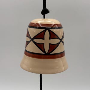 May include: A ceramic bell with a cream-colored glaze featuring a geometric pattern of brown and black lines. The bell hangs from a black cord, which also supports a matching circular ceramic clapper decorated with the same brown and black design.