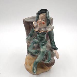 May include: A ceramic figurine vase featuring a seated elf character dressed in a green outfit with a dark green cap and a ruffled collar. The elf is perched on a brown, textured tree stump-shaped vessel with a hollow interior and a tan base.