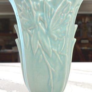 May include: A light blue ceramic vase with a glossy finish and a flared, scalloped rim. The body of the vase features a raised, embossed floral pattern with leaves and stems extending vertically along the sides, resting on a circular pedestal base.