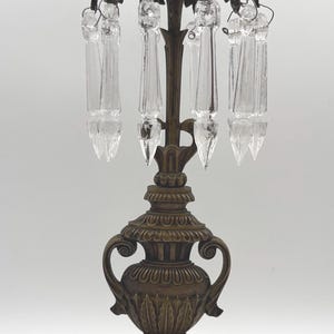 May include: A bronze-toned metal candelabra featuring a central urn-shaped base with ornate scroll handles, resting on a rectangular marble plinth. The top section displays hanging clear glass crystal prisms suspended from a decorative leaf-patterned rim.