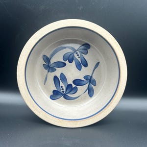 May include: A round, speckled stoneware bowl with a light beige base color. The interior features hand-painted blue floral motifs with three distinct petals and small dots. A thin blue circular band lines the inner rim of the bowl.