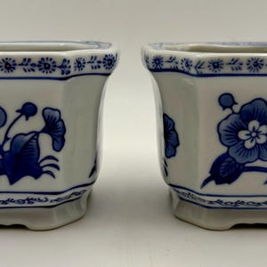 May include: Two white ceramic planters with a hexagonal shape and a blue floral pattern. Each planter features a decorative blue border along the top rim and base, with a central blue flower illustration on the front and side panels.