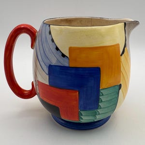 May include: A ceramic pitcher featuring a bright red handle and a colorful geometric pattern of yellow, blue, orange, green, and black shapes. The body of the pitcher has a rounded shape with a wide spout and a solid blue circular base.