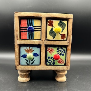 May include: A small wooden chest with four square drawers, each featuring a unique hand-painted design. The top left drawer has red and blue stripes, the top right has floral motifs, the bottom left has a blue flower, and the bottom right has red and blue patterns. Each drawer has a round, solid-colored ceramic knob.